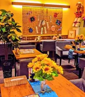 ワンぽてぃとの夏季装飾が施された明るい店内。黄色い壁、すだれ飾り、ひまわりの花が置かれたテーブル、グレーのソファ席が並ぶ温かみのある空間。