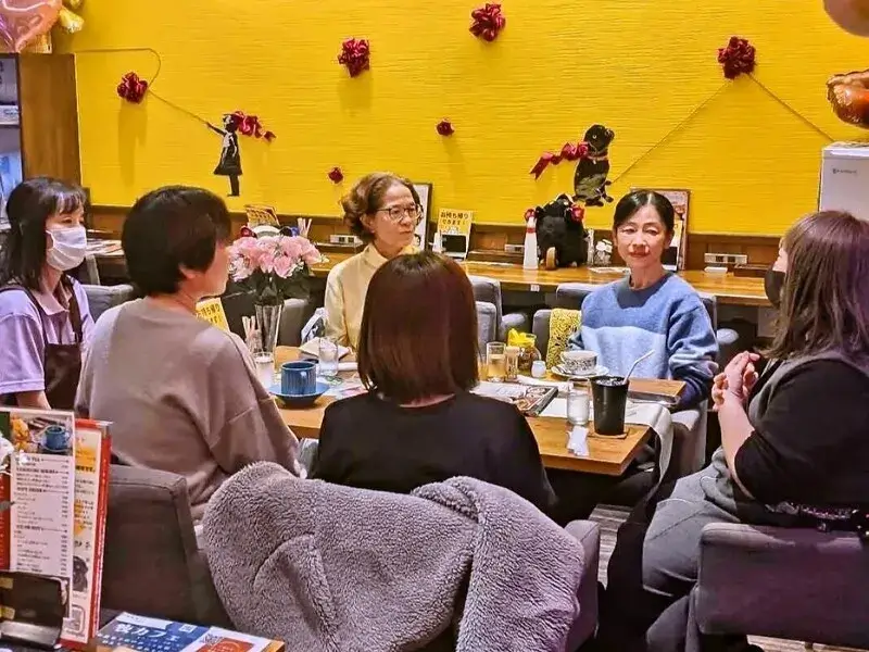 黄色い壁の飲食店で円卓を囲み、交流する女性たちのグループ。テーブルには飲み物や軽食が並んでいる。