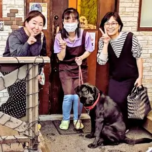 店の入り口前で笑顔でピースサインをする女性３人と、座っている黒いラブラドールレトリバー。