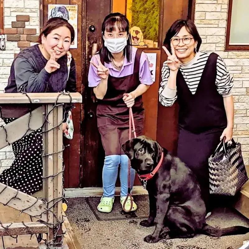 店の入り口前で笑顔でピースサインをする女性３人と、座っている黒いラブラドールレトリバー。