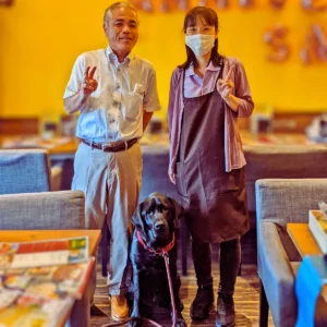 制度の狭間で苦しむ人々を救いたいと大阪から来店された男性のお客様と、ワンぽてぃとの店主、看板犬の黒いラブラドールレトリバー「ぽてぃと」の3ショット。笑顔でピースサインをする様子。