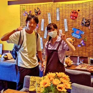 店員と笑顔でピースサインをする、日焼けした若い男性とマスク姿の女性。背景には「じゃがボナーラ」「じゃがドリア」などの手書きメニューがある。
