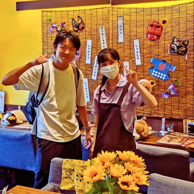 店員と笑顔でピースサインをする、日焼けした若い男性とマスク姿の女性。背景には「じゃがボナーラ」「じゃがドリア」などの手書きメニューがある。