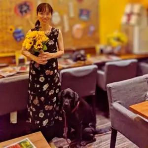 ひまわりの花束を抱えて優しい笑顔を見せる「ワンぽてぃと」のオーナー小栗加奈さん。その足元には、看板犬である黒いラブラドールのオリバーが寄り添って座っている。