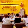 黄色い壁のカフェで、テーブルを囲んで和やかに話す5人の女性。壁には「HAPPY ANNIVERSARY」の文字バルーン装飾。