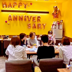 黄色い壁のカフェで、テーブルを囲んで和やかに話す5人の女性。壁には「HAPPY ANNIVERSARY」の文字バルーン装飾。