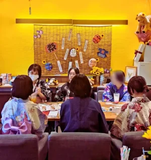 夏祭り風の飾りが施されたワンぽてぃと店内で、浴衣や甚平を着た主催者と若者たちがテーブルを囲む第3回HANASOU-ZEの様子。