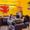 赤い着物が飾られたワンぽてぃと店内で、第8回HANASOU-ZEの参加者たちがテーブルを囲んでいる。向かい合って話す若者のうち、一人の女性が明るい笑顔を見せている。