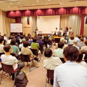 三重県松阪市のイベントで、ステージ上のパネリストの話に耳を傾ける満員の参加者。