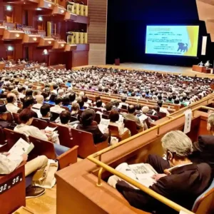 三重県民生委員・児童委員大会の講演会場。1000人以上の聴衆が、スクリーンと壇上の講演者を見ている様子。