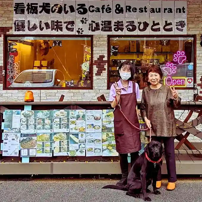 愛知県春日井市にあるカフェレストラン「ワンぽてぃと」の店先で、オーナーの小栗加奈さん(左の女性)と来店したお客様(右の女性)が、黒い看板犬と一緒にピースサインをして記念撮影している様子。