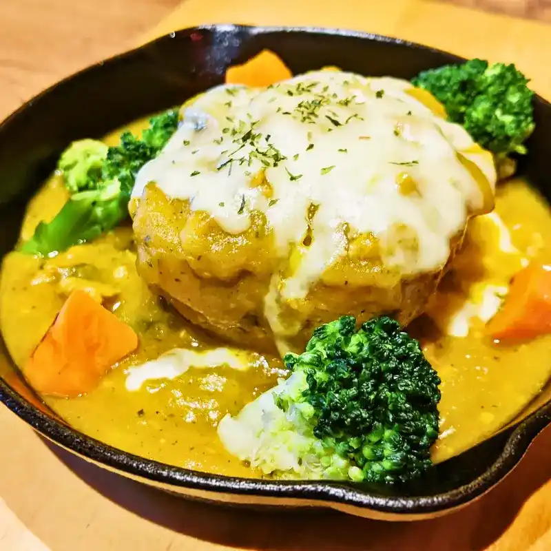 黒いスキレットに入ったOBAKEじゃがグラ。かぼちゃの黄色いソースの中に、チーズがおばけのように溶けたじゃがいもグラタンが置かれ、ブロッコリーが添えられている。