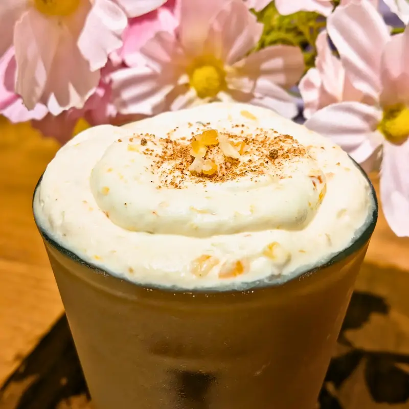 ホイップクリームがたっぷり乗った蜜芋オレンジオレ。オレンジピールとスパイスがトッピングされ、背景には花が飾られている。