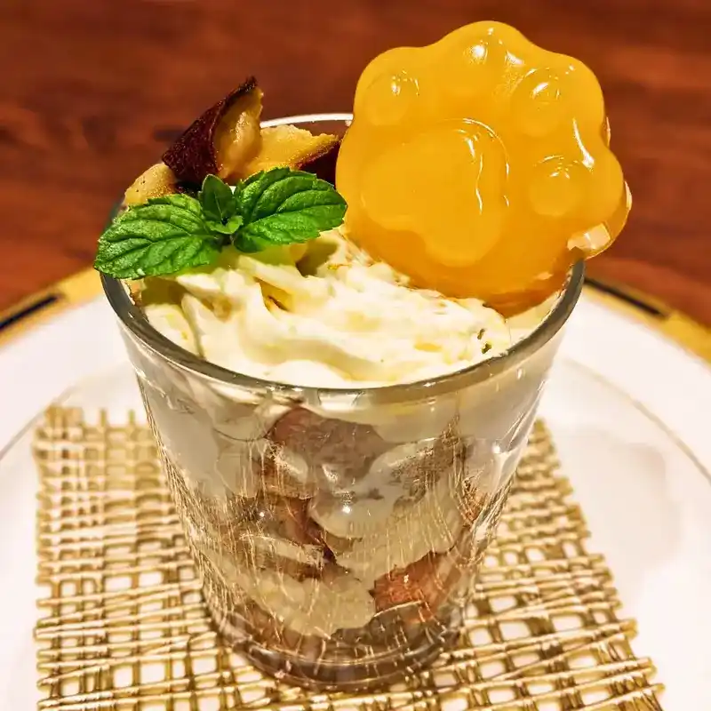 グラスに入ったオレンジと蜜芋のトライフル。層になったクリームとシフォンの上に、大きなオレンジ色の肉球ゼリーとミントが飾られている。