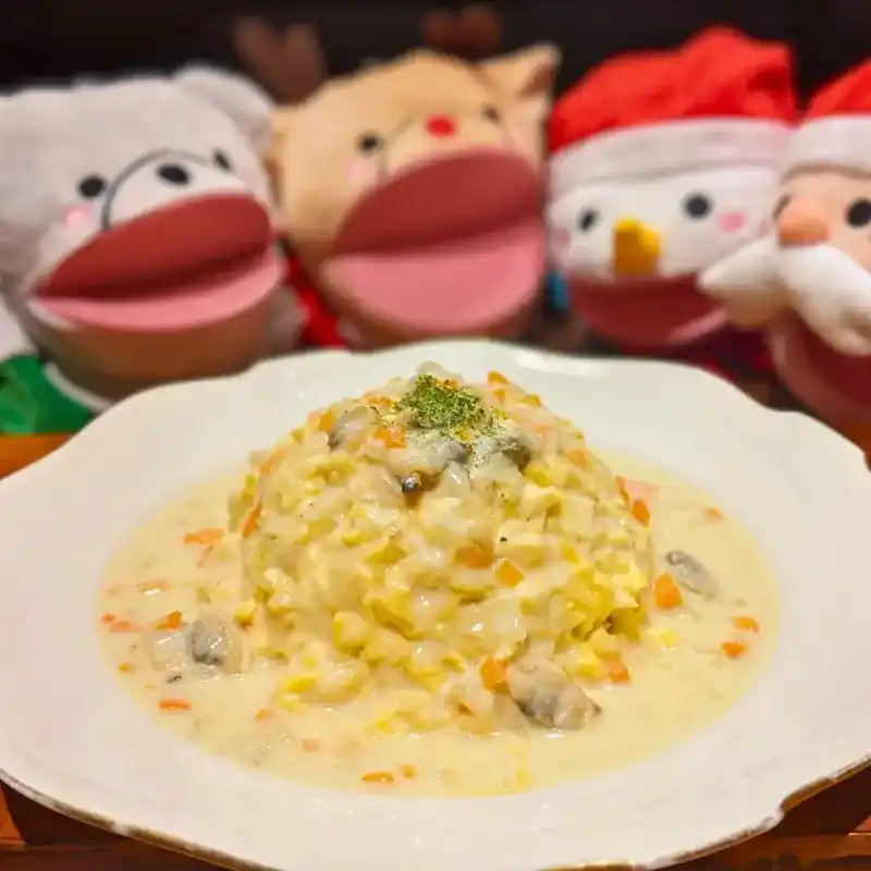 ふわふわの卵に白い濃厚なクラムチャウダーがたっぷりとかかった、クリスマス限定のホワイトオムライス。背景にはクリスマスのパペットが並んでいる。
