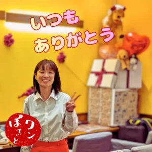 黄色い壁の明るい店内で、笑顔でピースサインをする「ワンぽてぃと」オーナーの小栗加奈。背景には「いつもありがとう」のメッセージ。