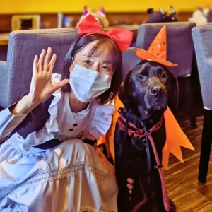 ハロウィンの魔女の帽子とマントをつけた黒ラブラドールの看板犬オリバーと、赤いリボンをつけ白いエプロンドレスを着たオーナーの小栗加奈が笑顔で並んでいる写真。