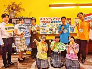 カフェレストラン「ワンぽてぃと」の店内で、手作りのフリップやマイクを持ってスピーチコンテスト用の動画撮影に取り組む春日丘高校の生徒たちと店主の集合写真