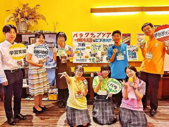 カフェレストラン「ワンぽてぃと」の店内で、手作りのフリップやマイクを持ってスピーチコンテスト用の動画撮影に取り組む春日丘高校の生徒たちと店主の集合写真