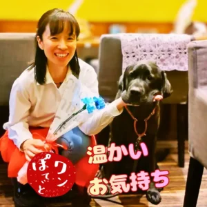 ワンぽてぃとの4周年記念にいただいた青いお花を手に持ち、看板犬の黒ラブラドール「オリバー」と笑顔で並ぶ小栗加奈の様子