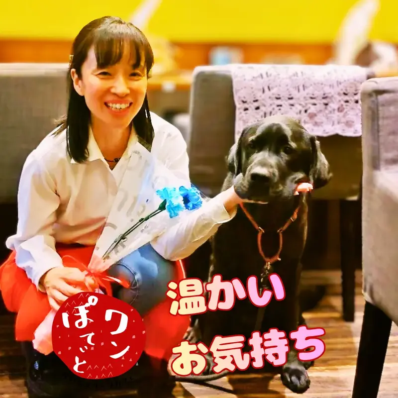 ワンぽてぃとの4周年記念にいただいた青いお花を手に持ち、看板犬の黒ラブラドール「オリバー」と笑顔で並ぶ小栗加奈の様子
