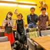 春日井市のカフェレストラン「ワンぽてぃと」で開催された5月のJAZZ LIVEにて。ライブのチラシを持つスタッフ、出演した女性ピアニスト率いるトリオバンドの皆様、そして手前に座る黒犬（オリバー）が並んで笑顔を見せている和やかな記念写真。