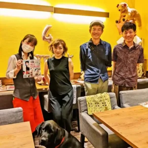 春日井市のカフェレストラン「ワンぽてぃと」で開催された5月のJAZZ LIVEにて。ライブのチラシを持つスタッフ、出演した女性ピアニスト率いるトリオバンドの皆様、そして手前に座る黒犬（オリバー）が並んで笑顔を見せている和やかな記念写真。