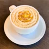 木目のテーブルに置かれた白いカップとソーサーに入ったホットの「蜜芋マロンオレ」。表面には渦巻き状のクリームがたっぷりと絞られ、中央に黄色いソースと刻んだ栗、茶色いパウダーがトッピングされている。