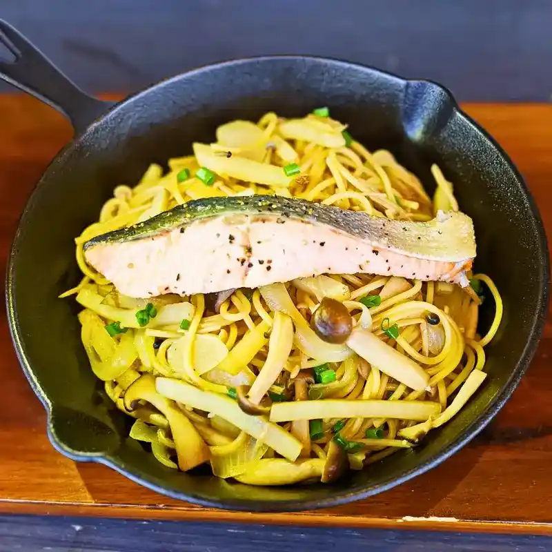 黒いスキレットに入った「焦がしバター醤油香る 鮭ときのこのじゃがパスタ」。細切りのじゃがいもを使ったパスタに、しめじやエリンギなどのきのこと小ねぎが絡み、中央に黒胡椒が振られた大きな焼き秋鮭の切り身が豪快にのっている。