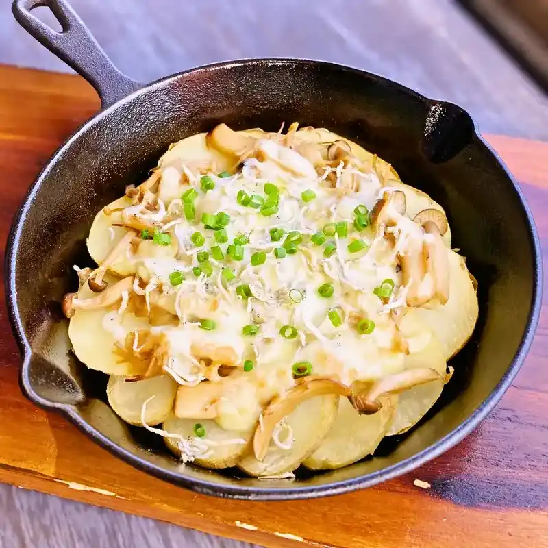黒いスキレットに盛り付けられた、輪切りのじゃがいもをベースにした「しらすときのこ 和風じゃがピザ」。とろけるチーズの上に、たっぷりのしらすと、しめじやエリンギなどのきのこ、彩りの小ねぎが散らされている。