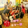 春日井市のカフェレストラン「ワンぽてぃと」店内でオーナーの小栗加奈、看板犬オリバー、中部中学校ギター部の顧問と生徒が楽器を持って笑顔で並ぶ集合写真
