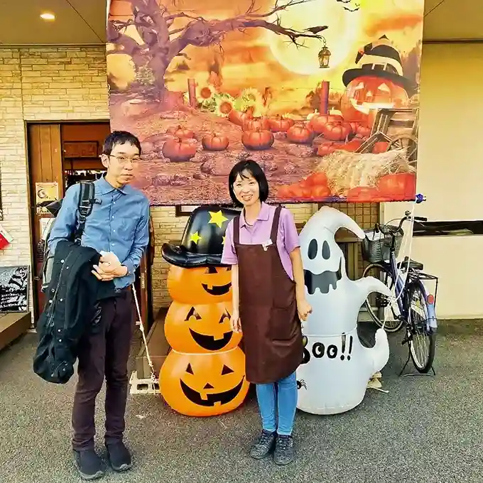 カフェレストラン「ワンぽてぃと」の店先で、ジャック・オー・ランタンやおばけのバルーンなど賑やかなハロウィンの飾り付けを背景に、笑顔で並ぶオーナーの小栗加奈と愛知教育大学の川北先生。
