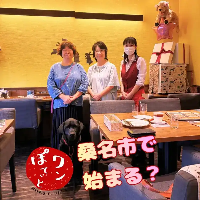 カフェレストラン「ワンぽてぃと」の店内で笑顔で並ぶ、三重県桑名市のひきこもりセンタースタッフとオーナーの小栗加奈、手前にお座りする黒ラブの看板犬オリバー。